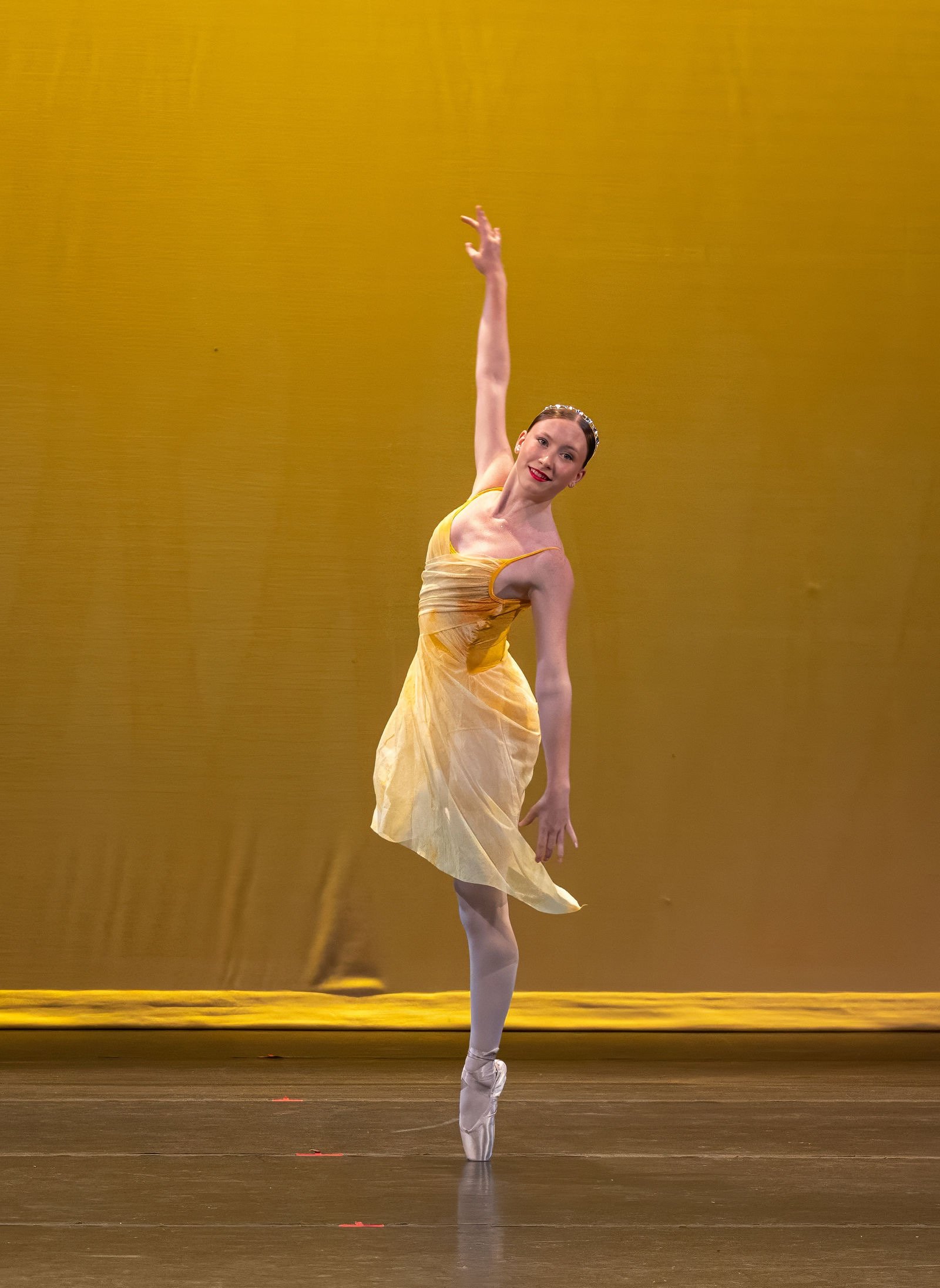 Ballet dancer on stage in a yellow dress performing en pointe with one arm raised and smiling