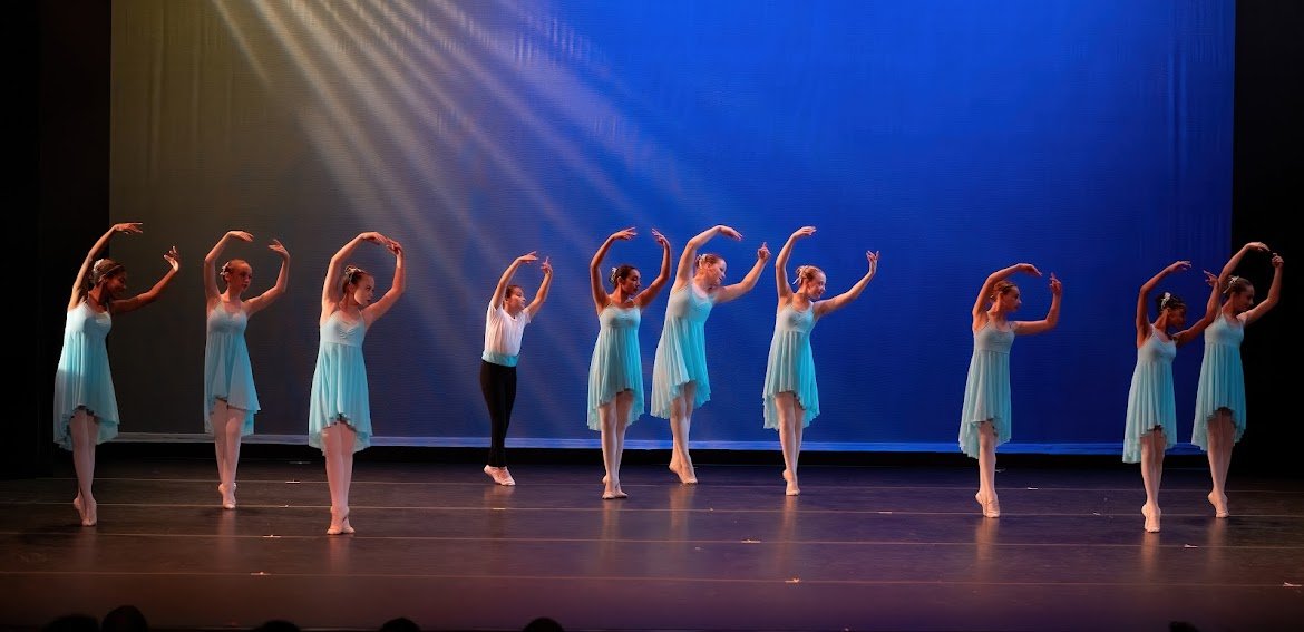 Ten ballet dancers in light blue dresses perform on stage with a dark background and a blue backdrop, with some dancers raising their arms above their heads.