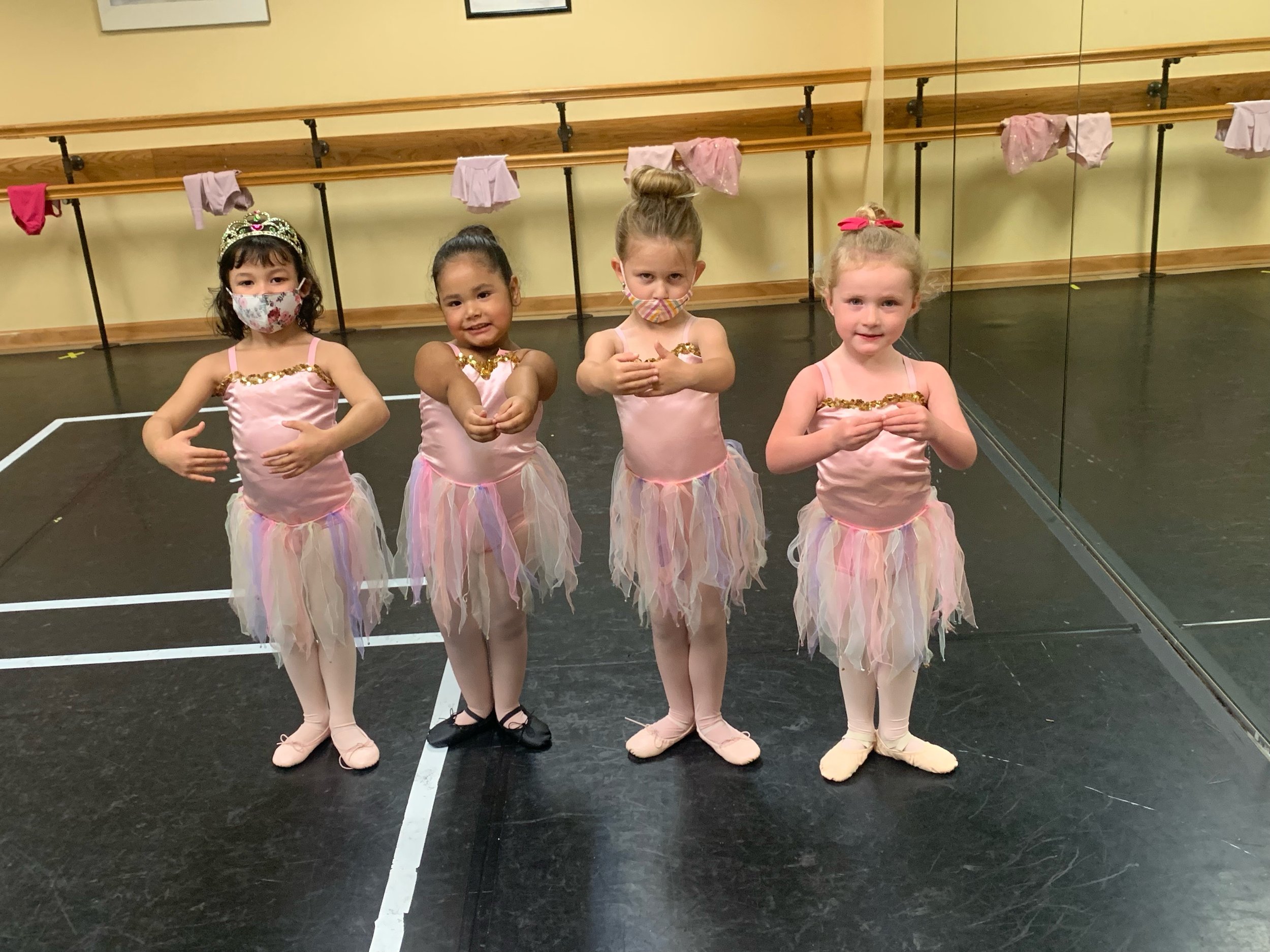 Four young girls dressed in pink ballet costumes with pastel tutus and ballet slippers, standing in a dance studio with a mirror, ballet barres, and pink clothing hanging on a wall.