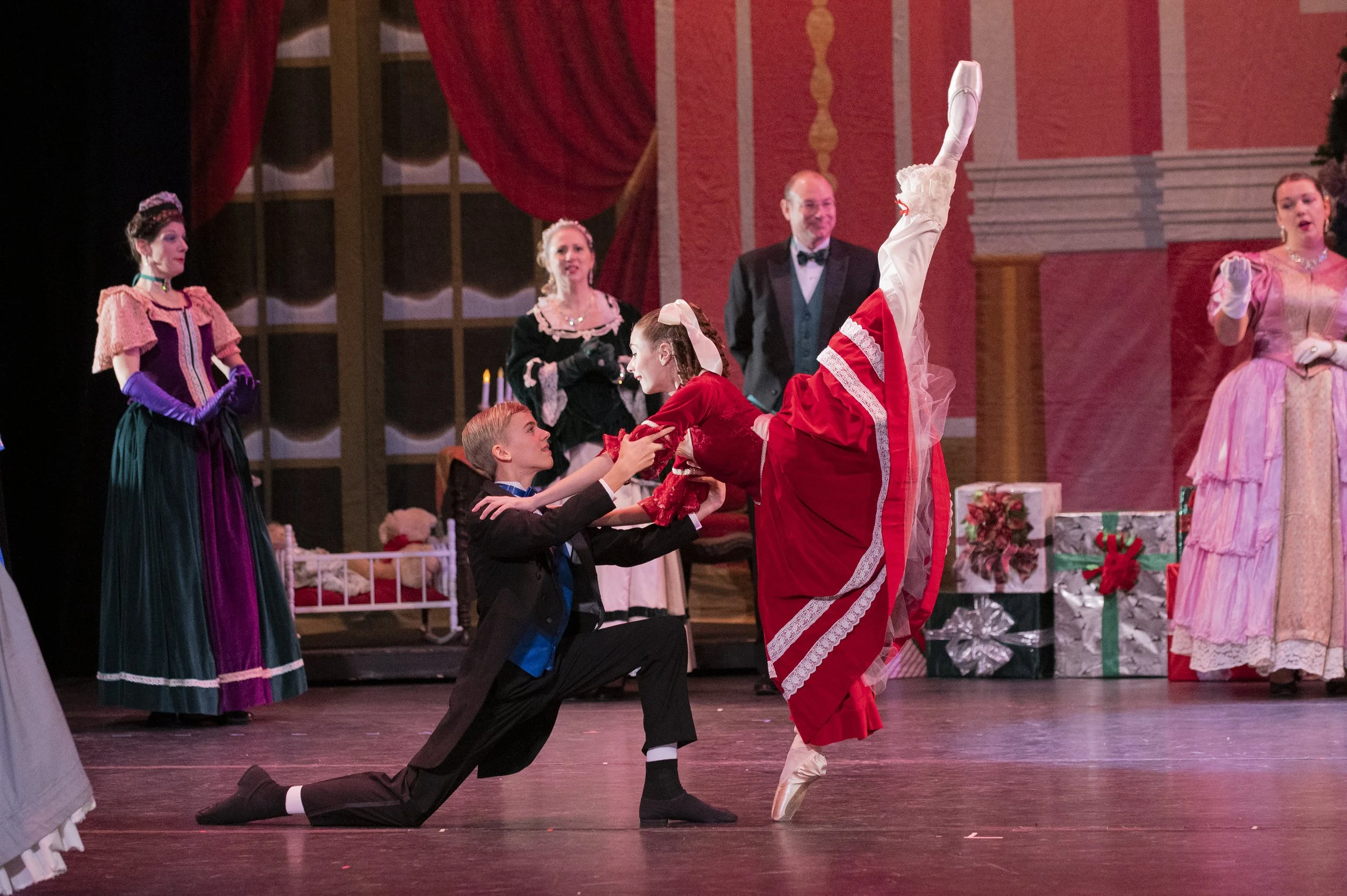 A ballet scene with a woman in a red costume performing an arabesque on pointe, while a man in a tuxedo kneels and holds her hands. Other women in period costumes and a man in a tuxedo stand in the background, watching in a decorated stage with large