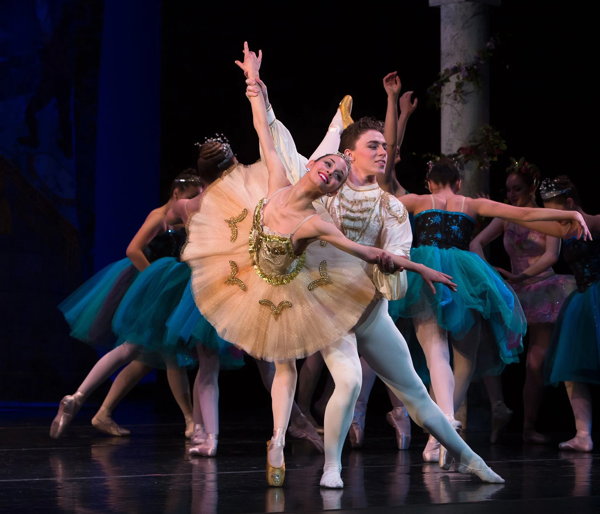 Ballet dancers performing on stage, with female dancers in tutus and male dancers in costumes, some with arms raised and expressions of concentration and grace.