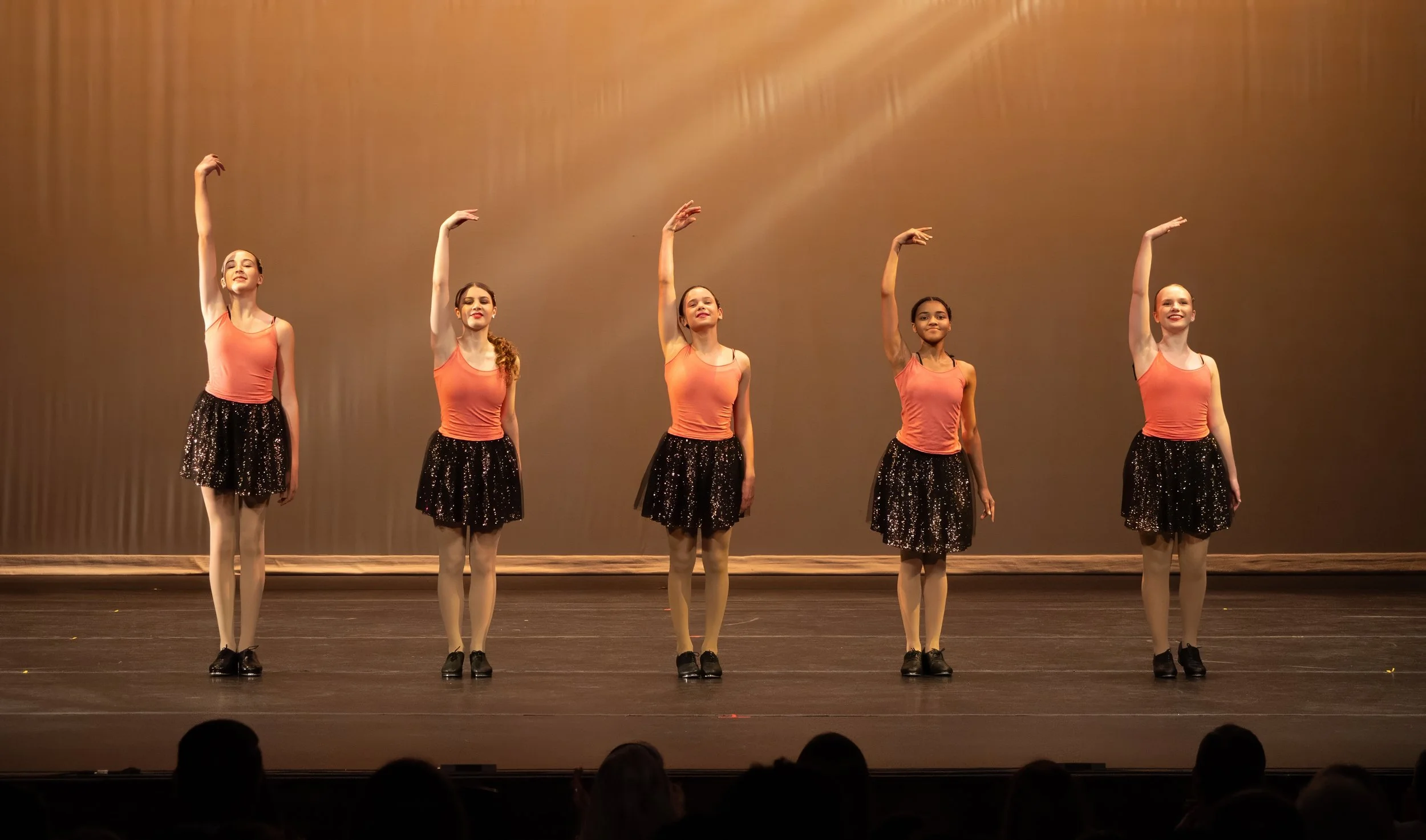 Five young ballet dancers in peach tops and black sparkly skirts standing on stage, with their right arms raised and feet together, performing a dance routine under warm stage lighting.