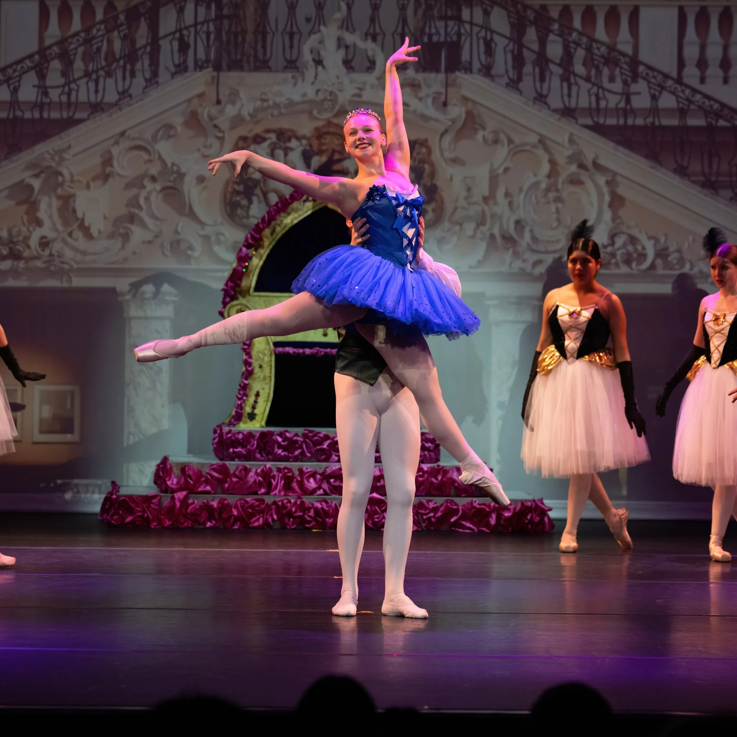 Ballet dancers performing on stage, with one dancer being lifted in a classical ballet pose, wearing a blue tutu, and others in pink tutus with black and gold accents standing in the background.