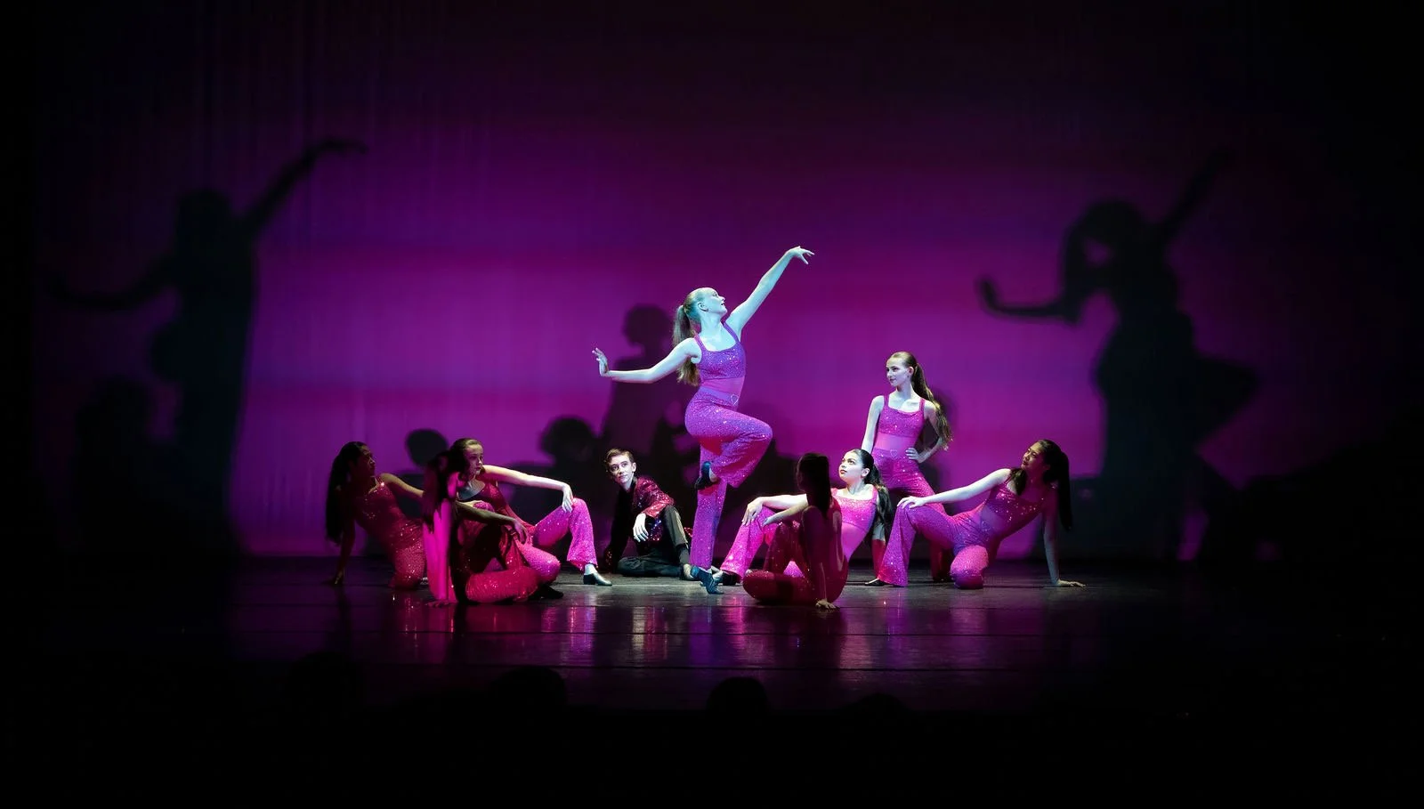 Group of young dancers on stage wearing pink costumes, performing a dance routine with dramatic shadows cast on the backdrop.