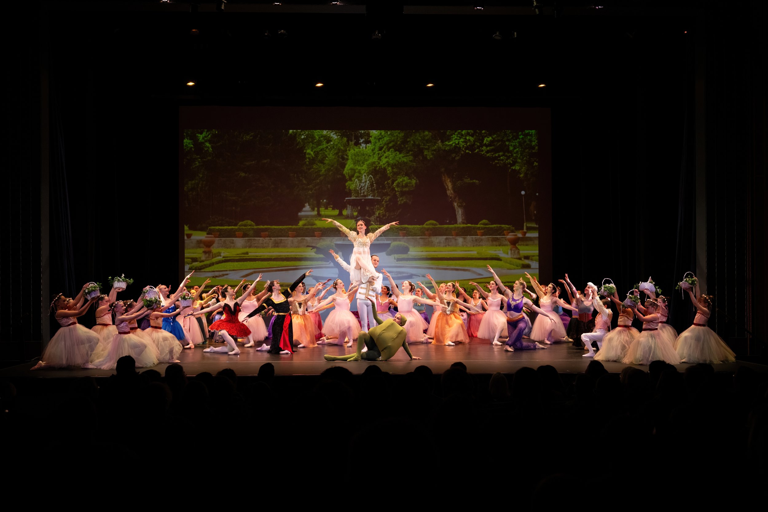Ballet dancers performing on stage with a scenic backdrop of a garden and fountain, with dancers in colorful costumes, some holding baskets of flowers, and a performer lifted in the center.