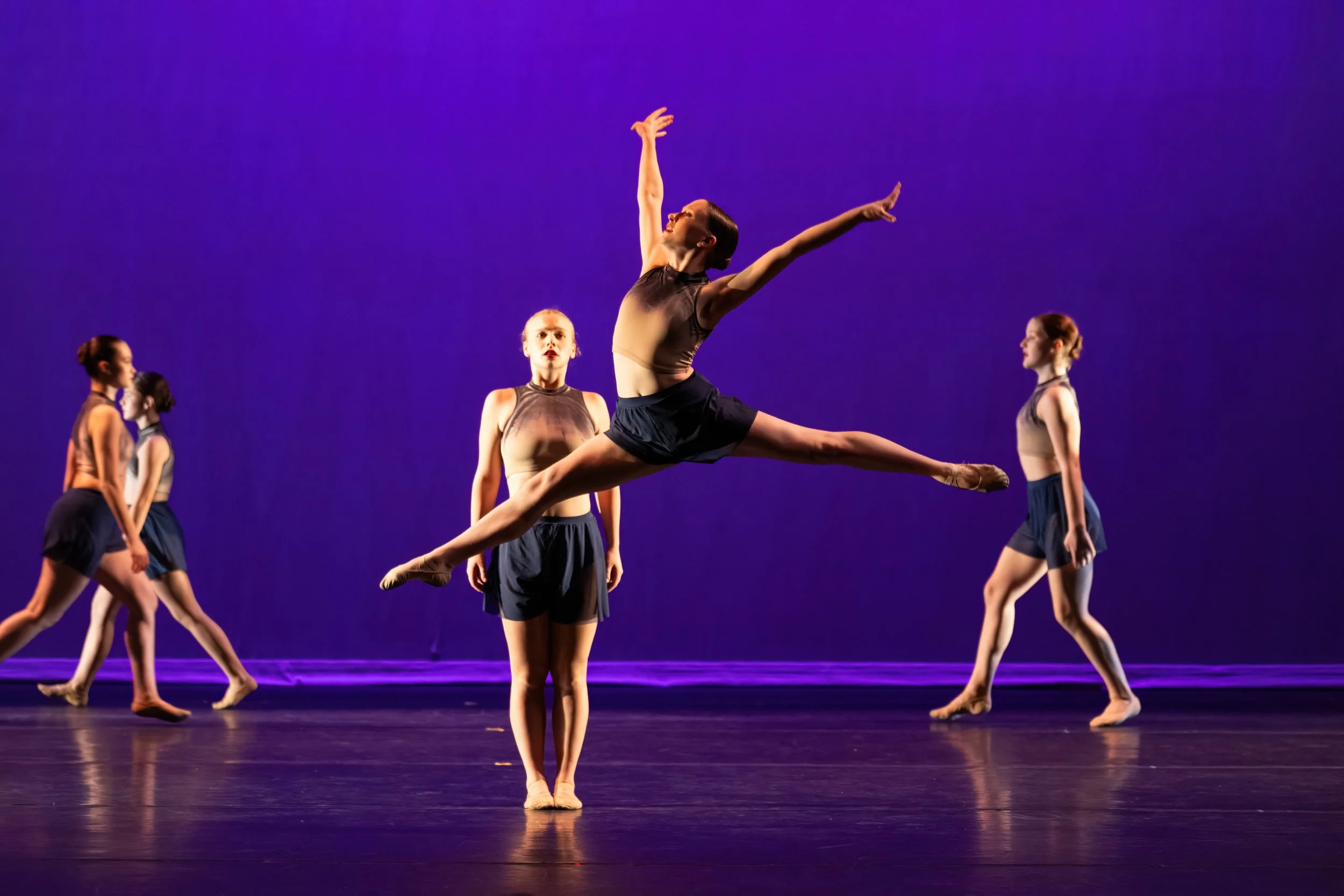 A group of ballet dancers performing on stage with a purple backdrop. One dancer is jumping in the air with legs extended and arms raised, while the others watch or pose in the background.