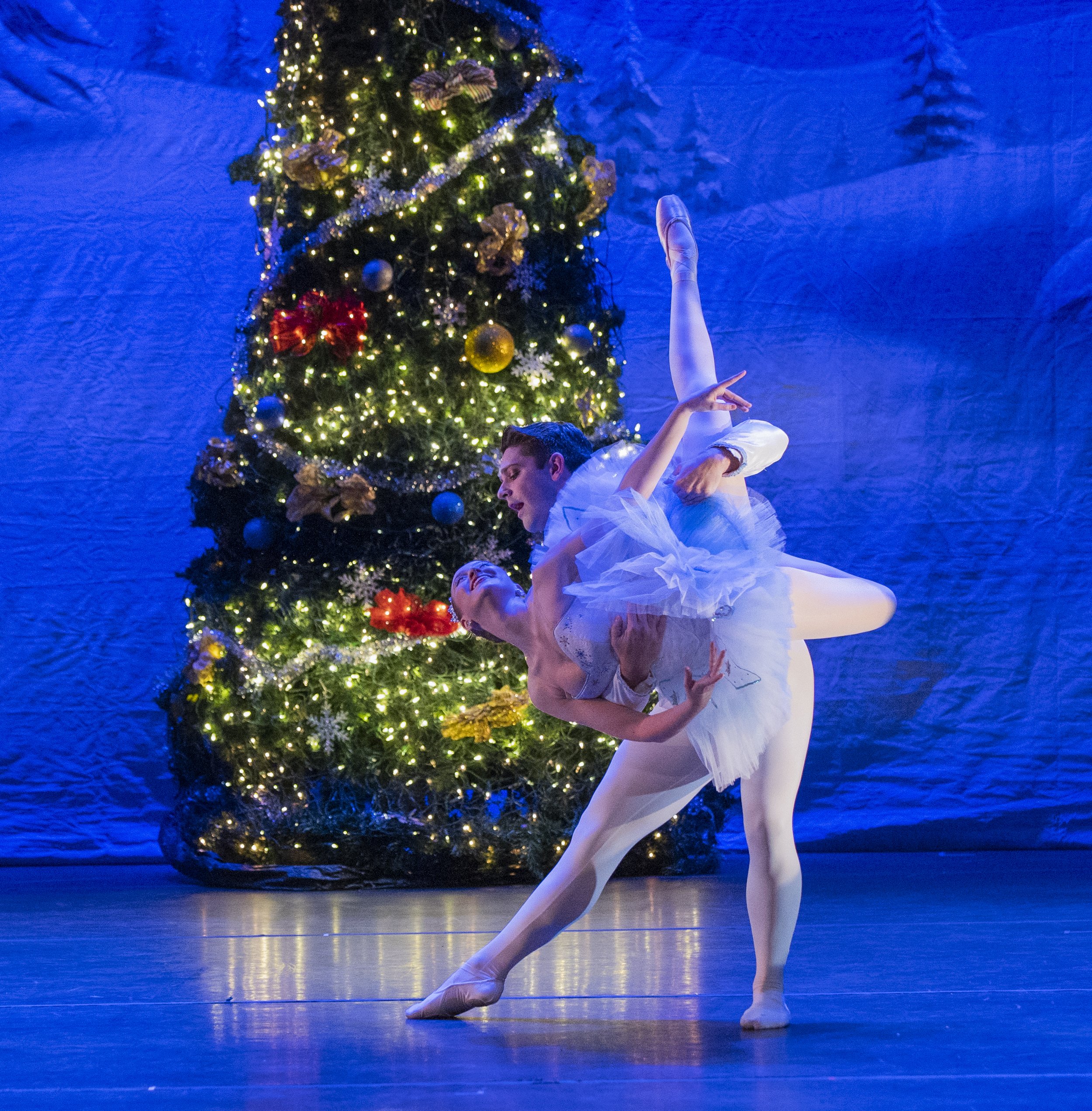 Two ballet dancers performing on stage with a decorated Christmas tree in the background, illuminated with colorful lights and ornaments.