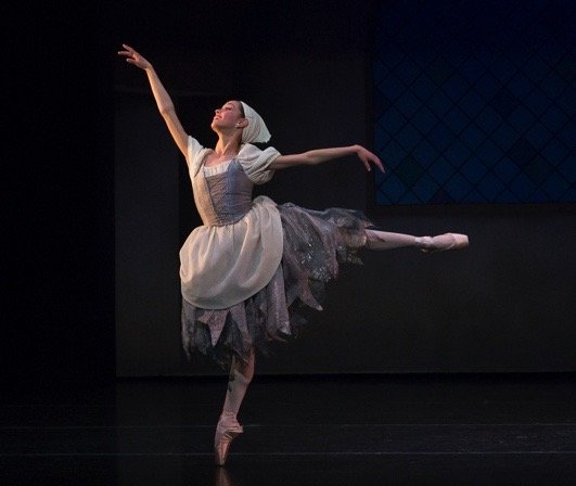 Ballet dancer performing on stage, wearing a white dress with a tulle skirt, balancing on one foot with arms extended gracefully.