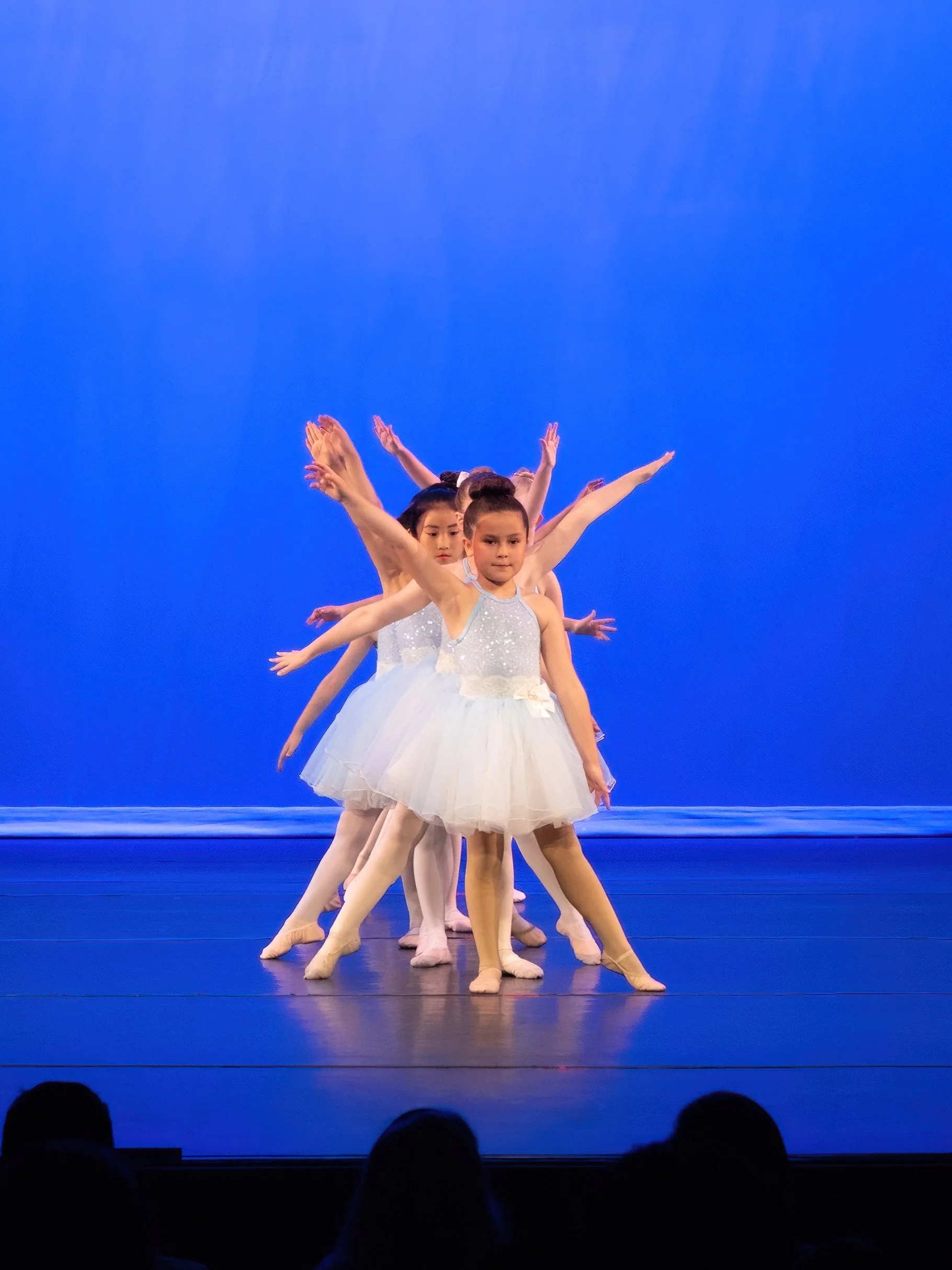 Young ballet dancers performing on stage in white tutus against a blue background.