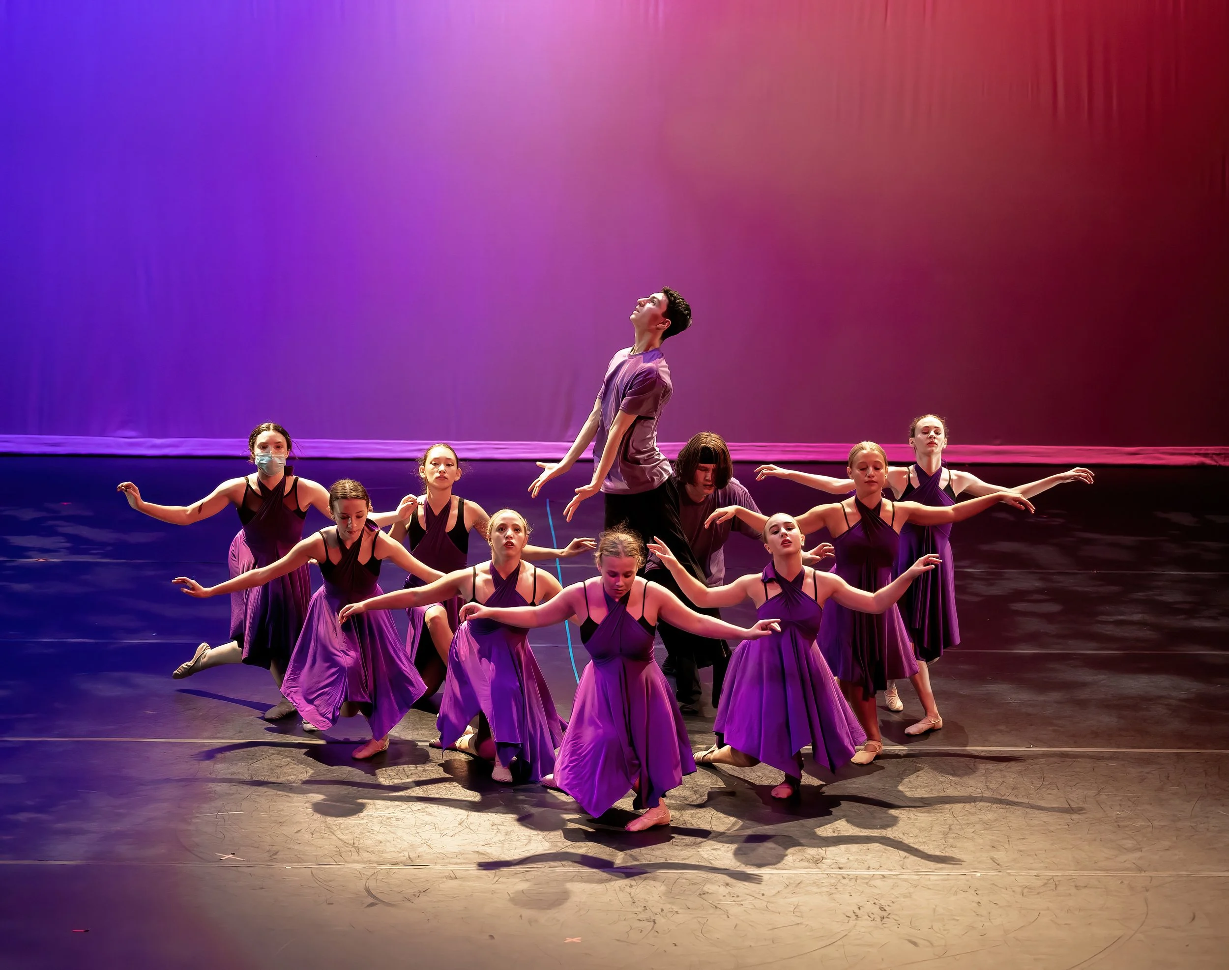 A group of young female dancers dressed in purple costumes performing a dance on stage with colorful lighting.