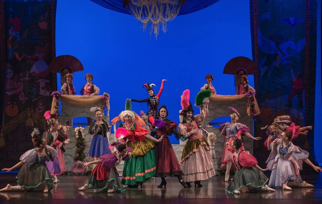 Theater scene with dancers in colorful costumes performing on stage with a blue backdrop, some with feathered accessories and props.