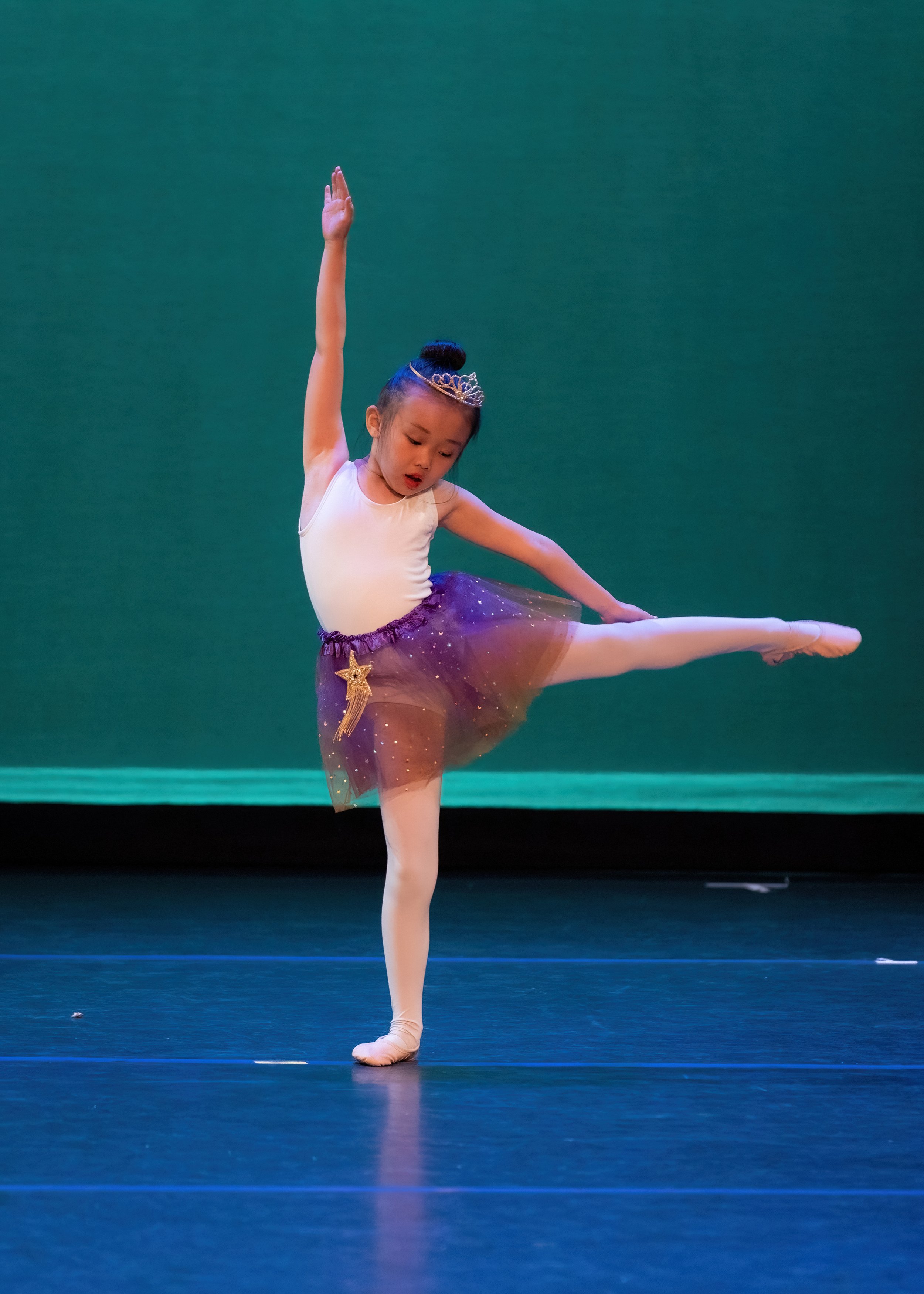A young girl performing a ballet dance on stage, wearing a white leotard, a purple tutu with a star and comet embellishment, a tiara, and ballet slippers.