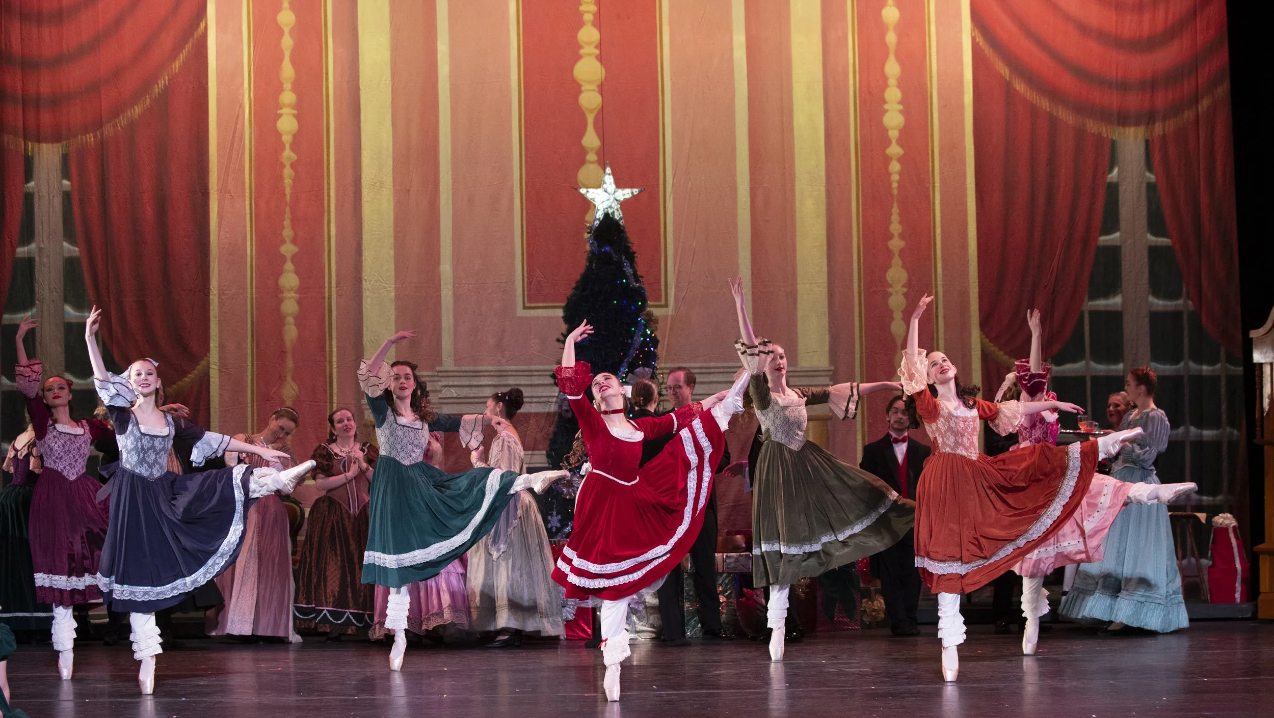 Ballet dancers in colorful costumes performing on stage with a Christmas tree and red curtains in the background.