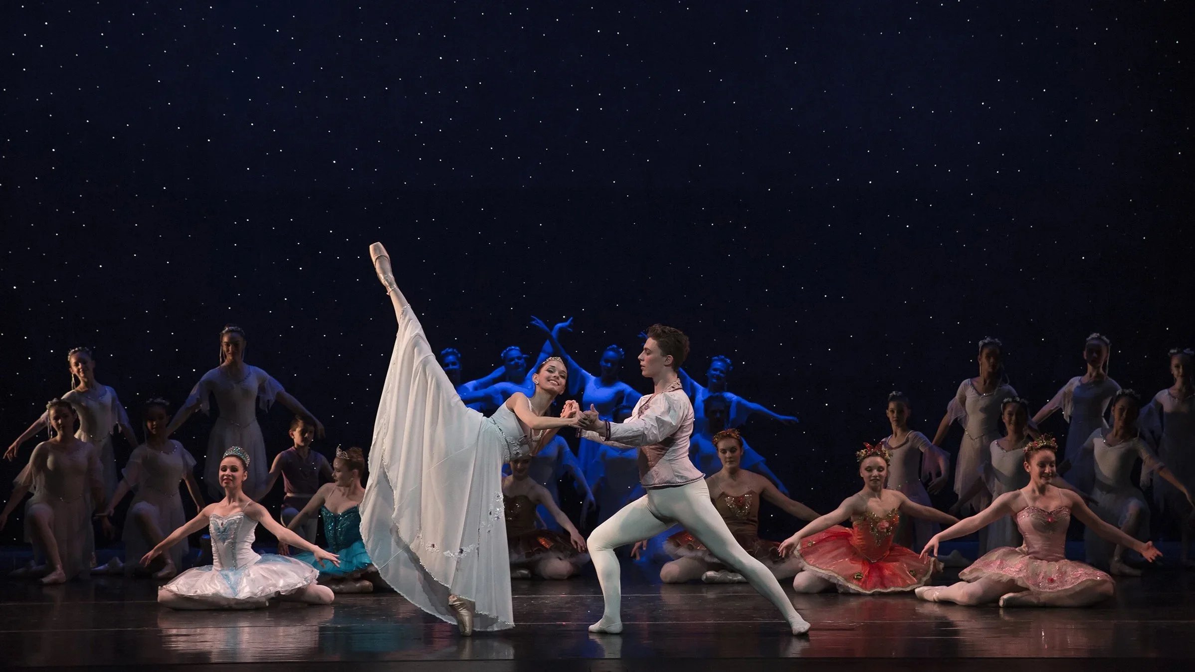 Ballet dancers on stage, with two principal dancers in the foreground performing a duet, surrounded by other dancers in colorful costumes, under a starry backdrop.