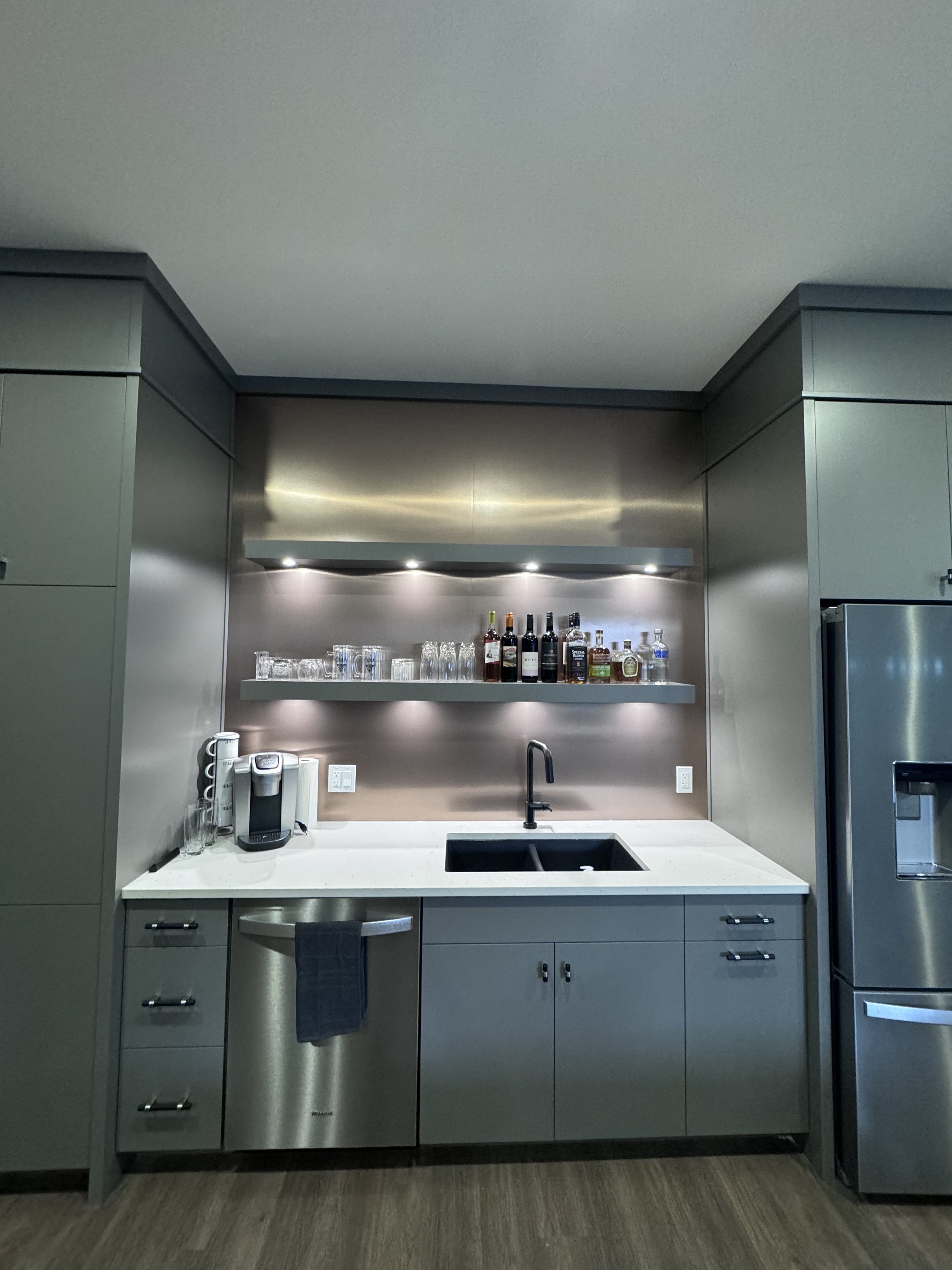 Modern kitchen with gray cabinets, a white countertop, a black sink, a coffee maker, and a shelf with liquor bottles and glasses, stainless steel appliances.