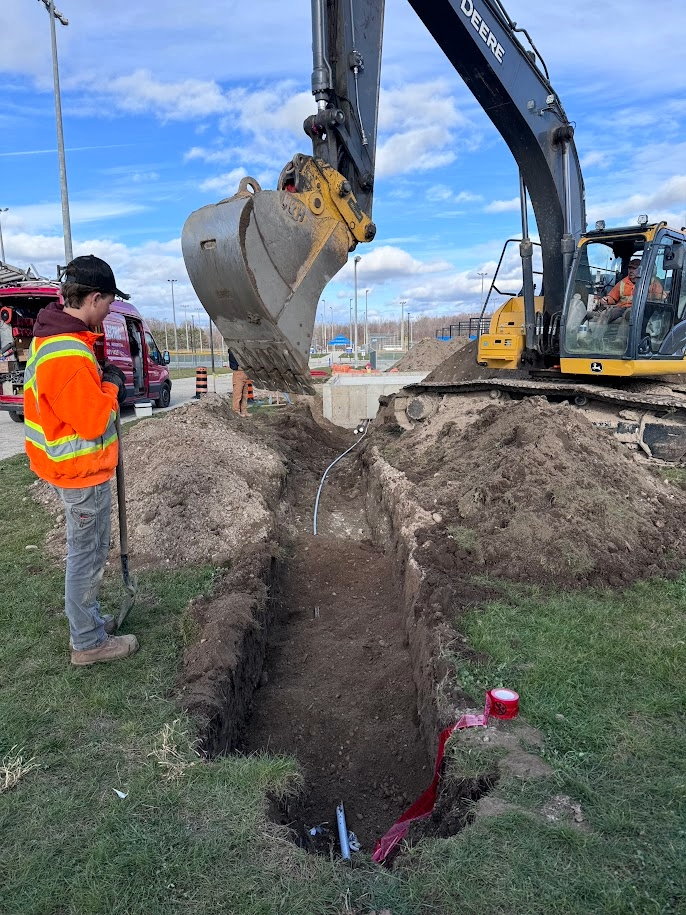 Construction site with an excavator digging a trench, with a worker in a high-visibility orange jacket and a worker in the excavator cab.