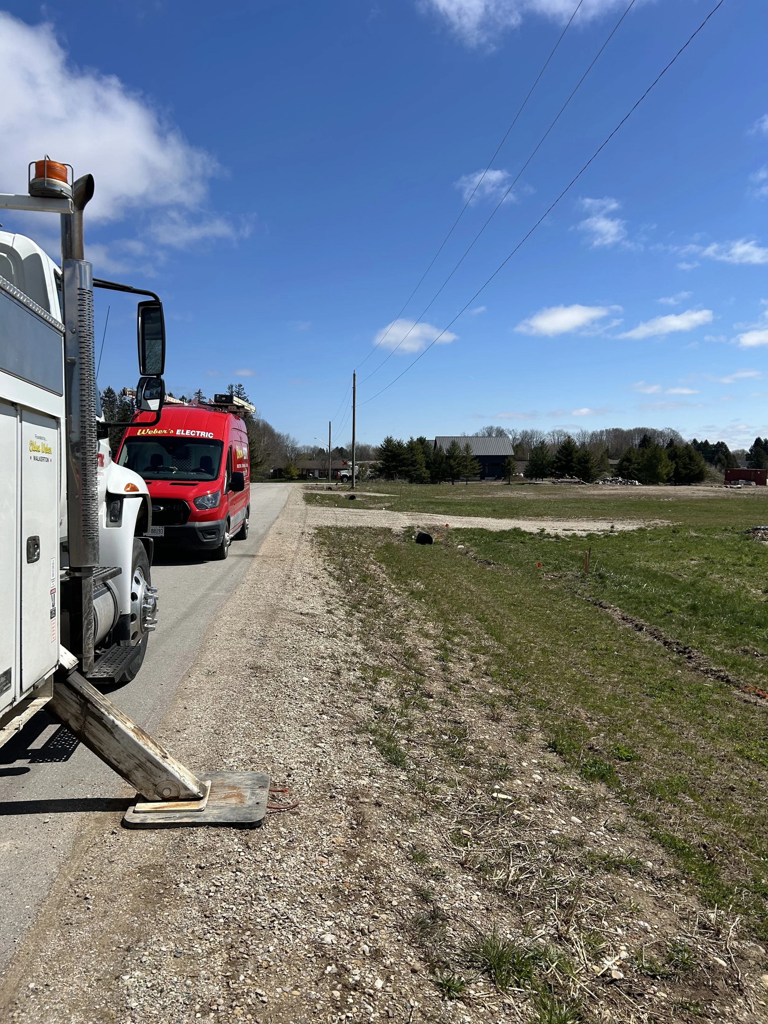 Vehicles parked on the side of a rural road with a gravel shoulder, and utility poles with power lines against a blue sky with scattered clouds. There are trees and a building in the distance.