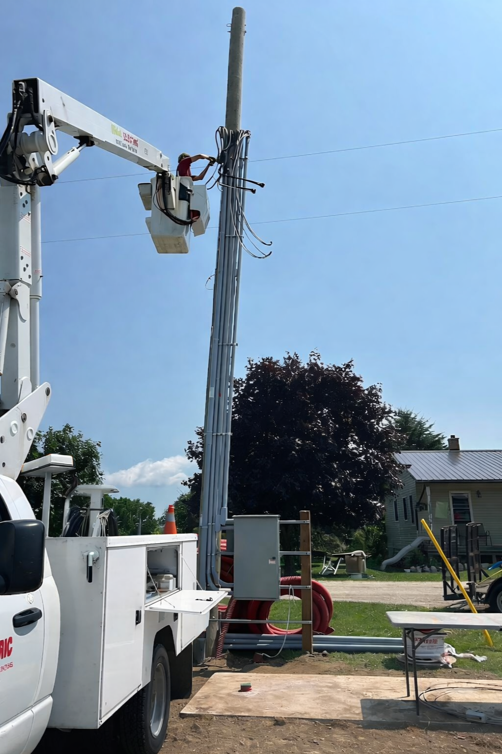 A worker in a bucket lift working on electrical wires on a utility pole during daytime. The utility truck is parked nearby on a grassy area, with trees, a house, and a playground in the background.