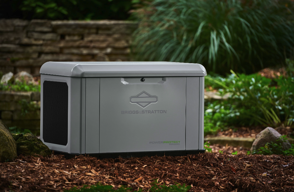 A gray Briggs & Stratton standby generator with a logo, situated in a garden with plants, rocks, and a stone wall in the background.