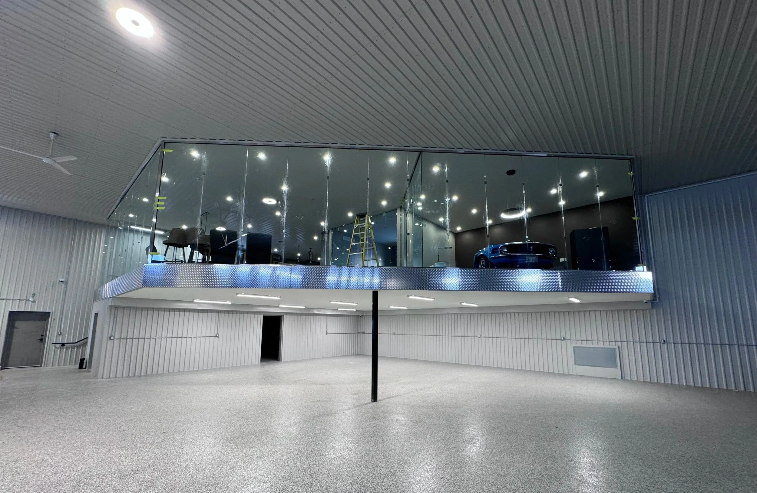 Large empty indoor space with a second-floor glass enclosure displaying a car. The room has a gray speckled floor, metal-paneled walls, and a ceiling with built-in lights.