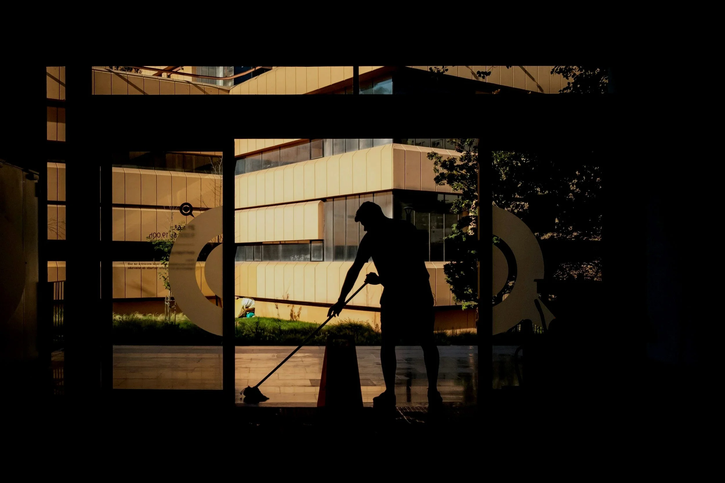Silhouette of a person cleaning the floor inside a building with large glass windows, showing a modern building and a bicycle outside.
