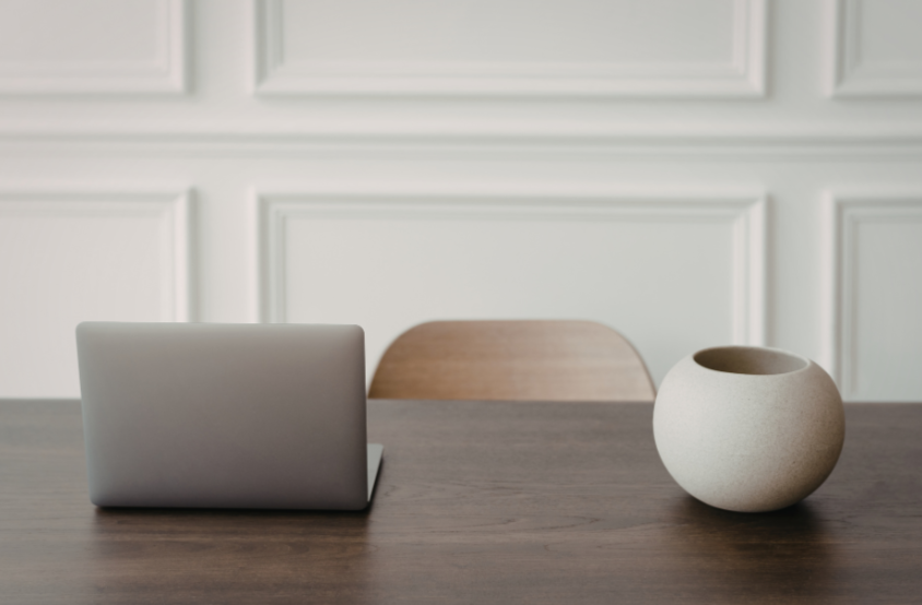 A minimalist desk setup with a closed silver laptop, a wooden chair, and a round beige ceramic vase on a dark brown wooden table in front of white paneled walls.