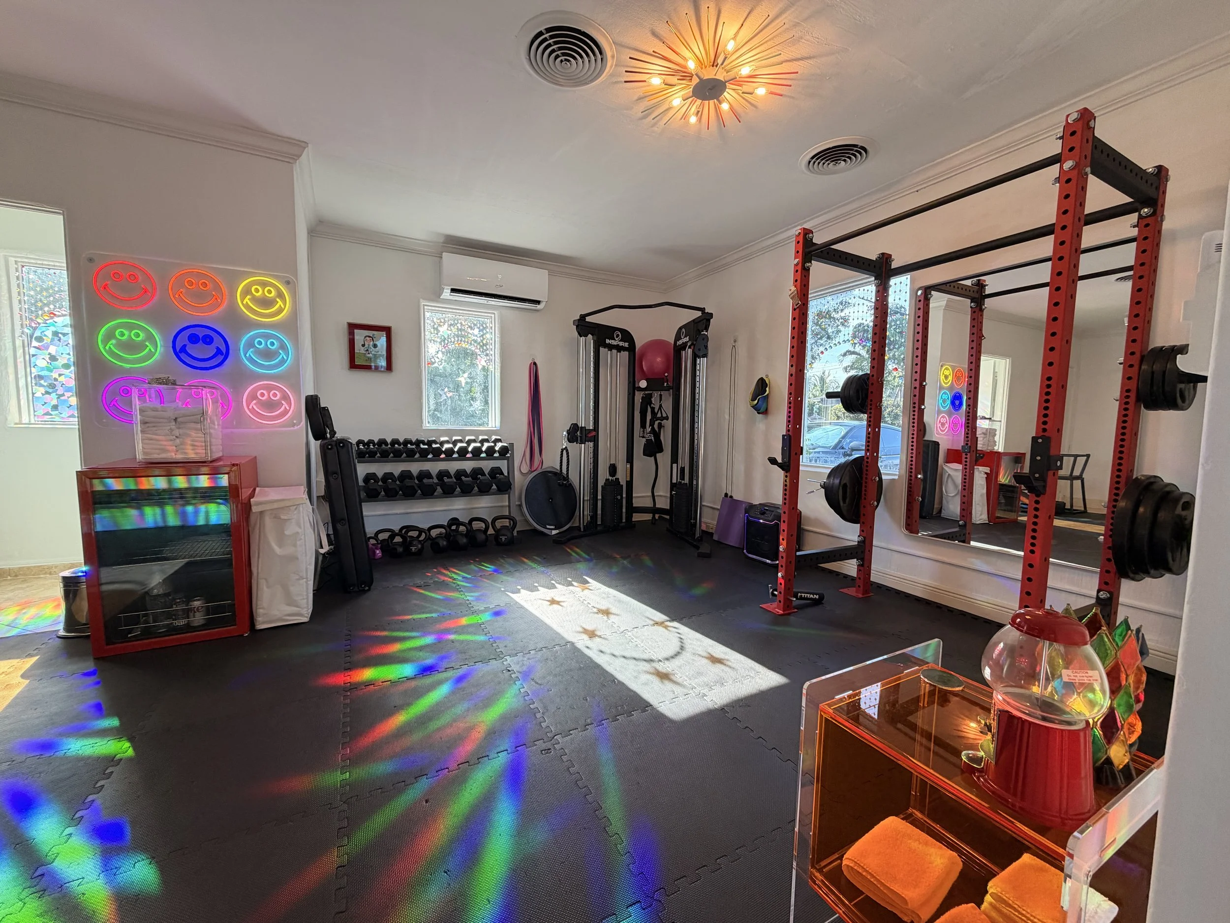Warm toned studio with workout equipment, neon smiley face lights on the wall, matted floors, and sunlight creating rainbow patterns on the floor.