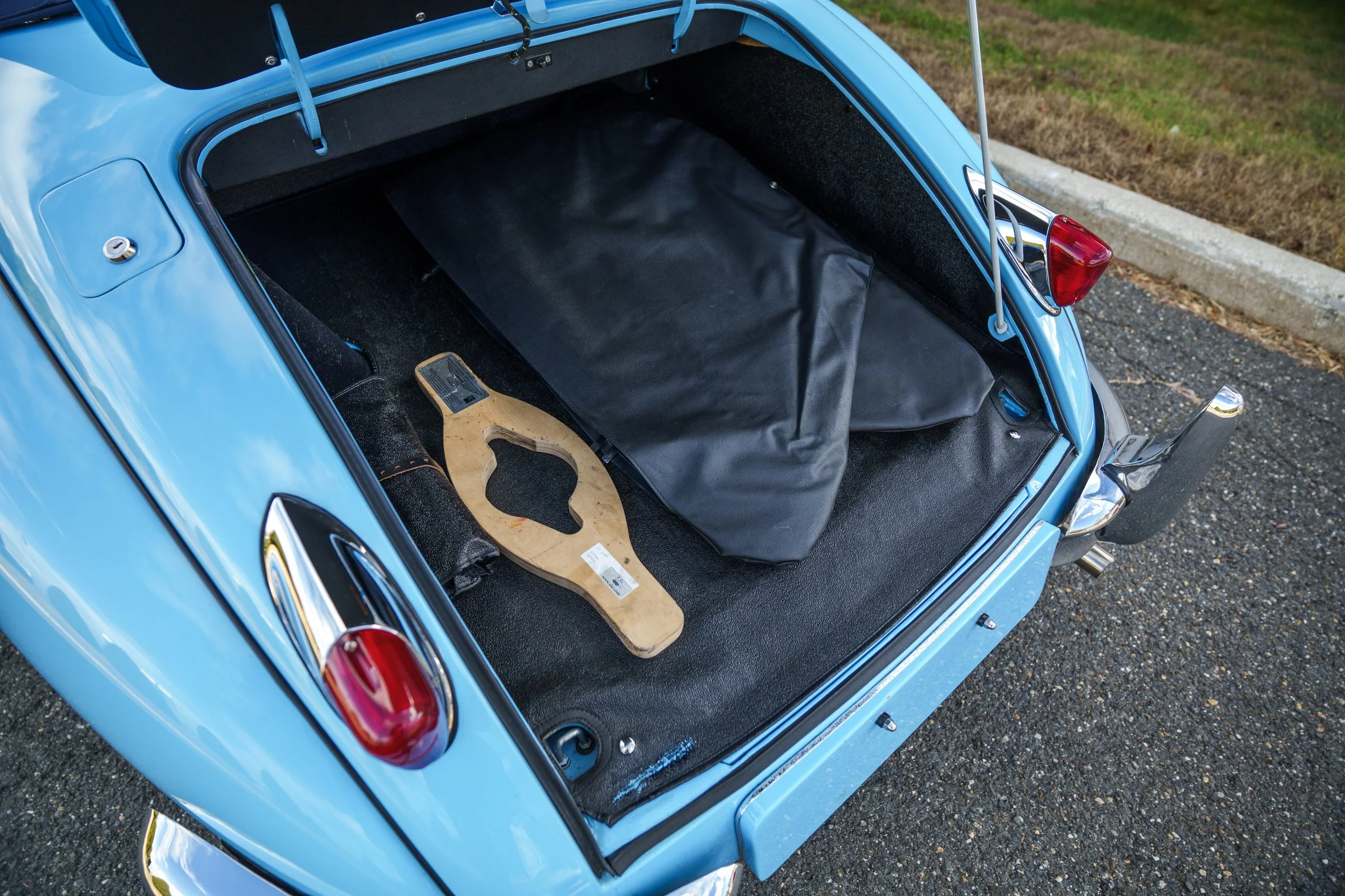 The image shows the open rear trunk of a vintage blue car with a black interior lining. Inside, there is a black bag and a wooden object that looks like a gasket or a frame. The car is parked on an asphalt surface next to a grassy area.