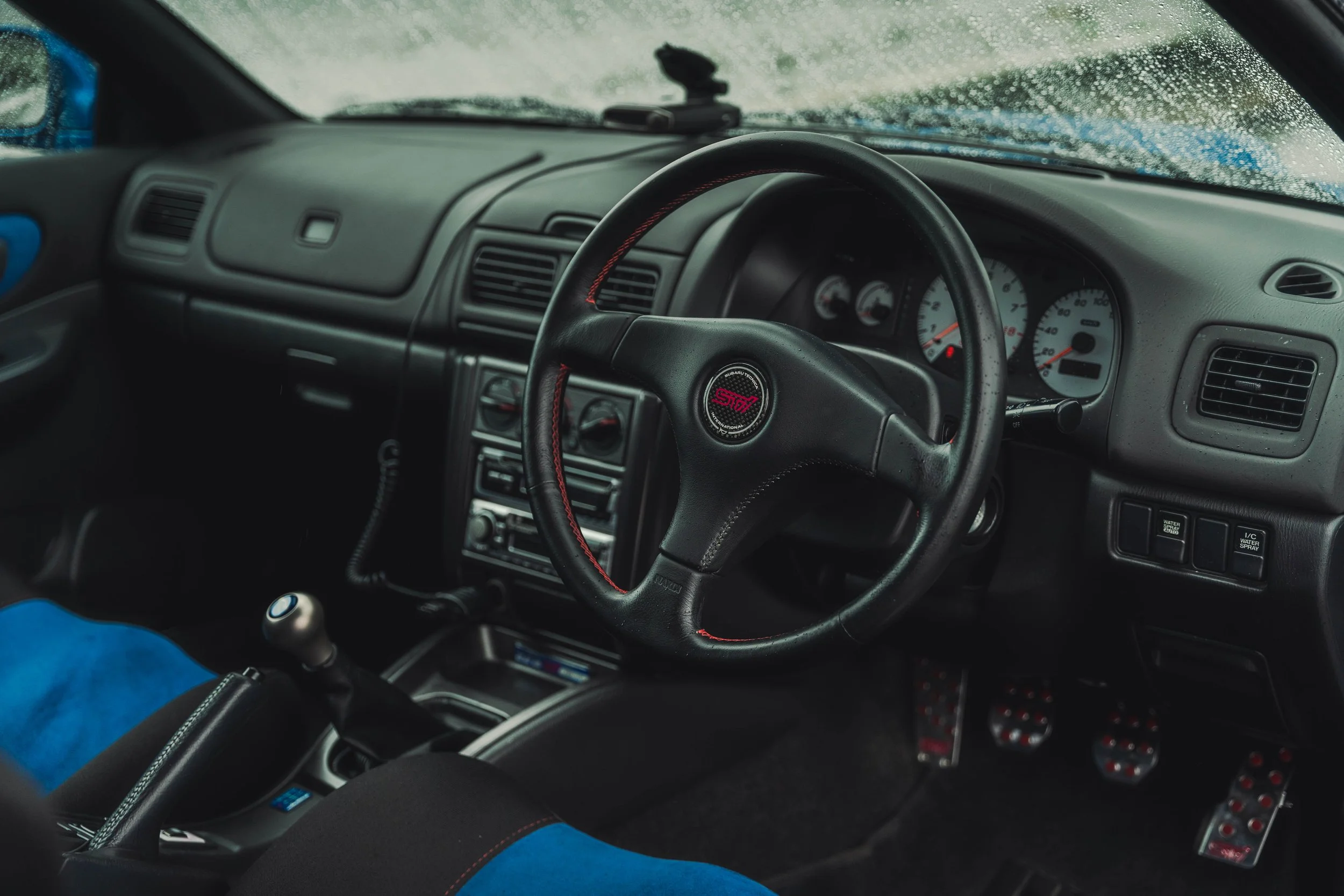 Interior view of a car with a steering wheel featuring STI logo, manual gear shift, and racing pedals.