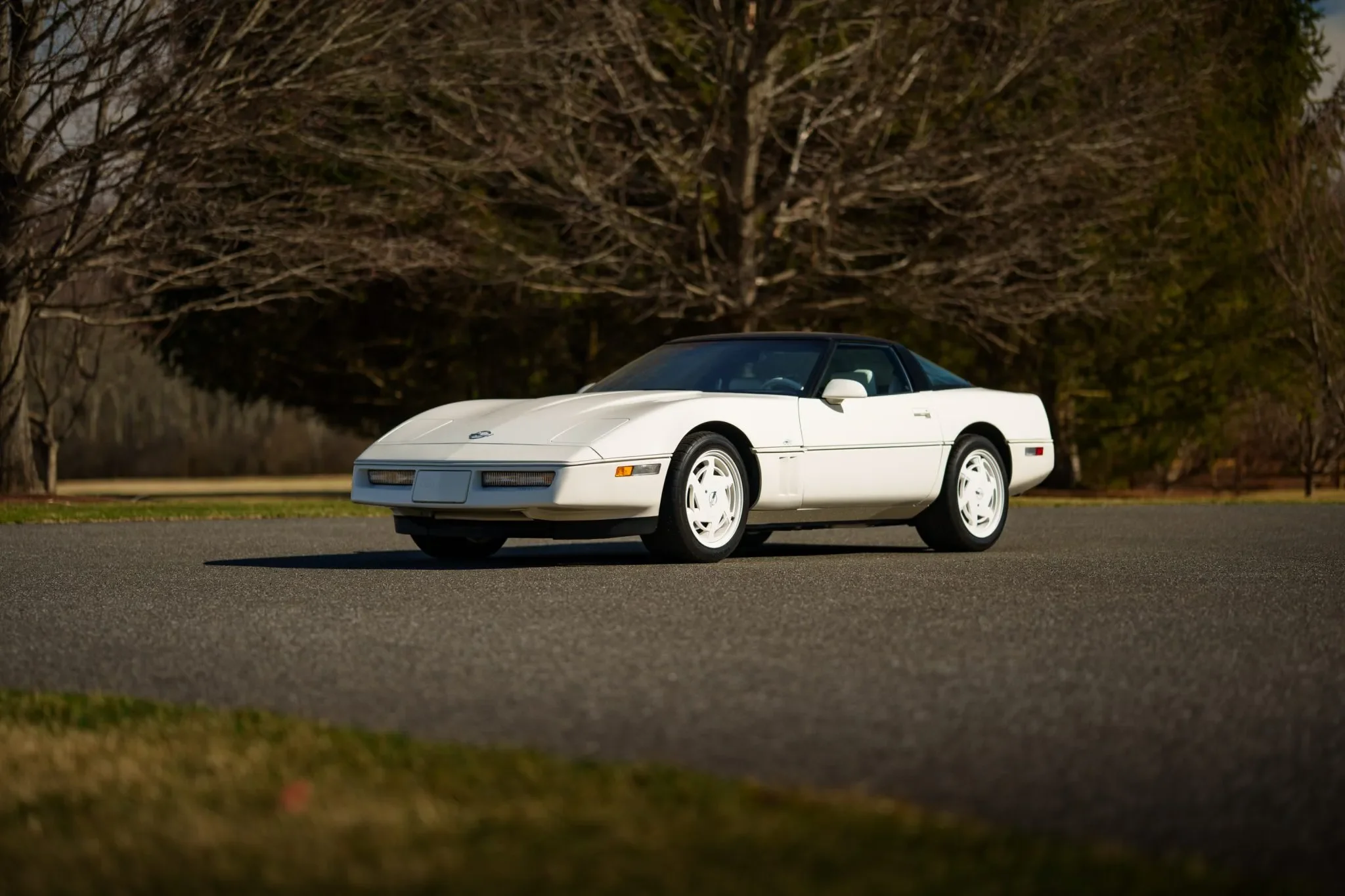 A white vintage sports car, possibly a Chevrolet Corvette, parked on a paved road with leafless trees in the background.