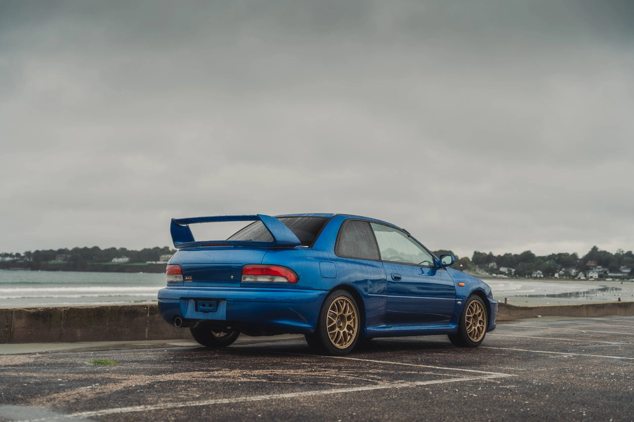 Blue car with a rear spoiler parked on a wet road near a body of water on a cloudy day.