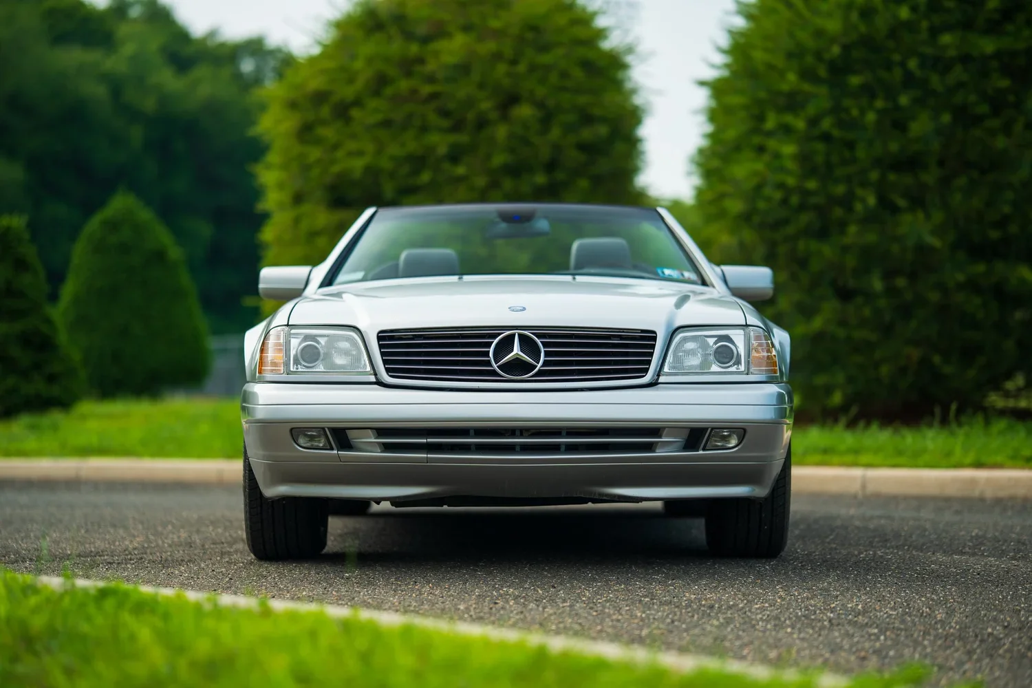Front view of a silver Mercedes-Benz convertible car parked on a paved asphalt surface with green trees and bushes in the background.
