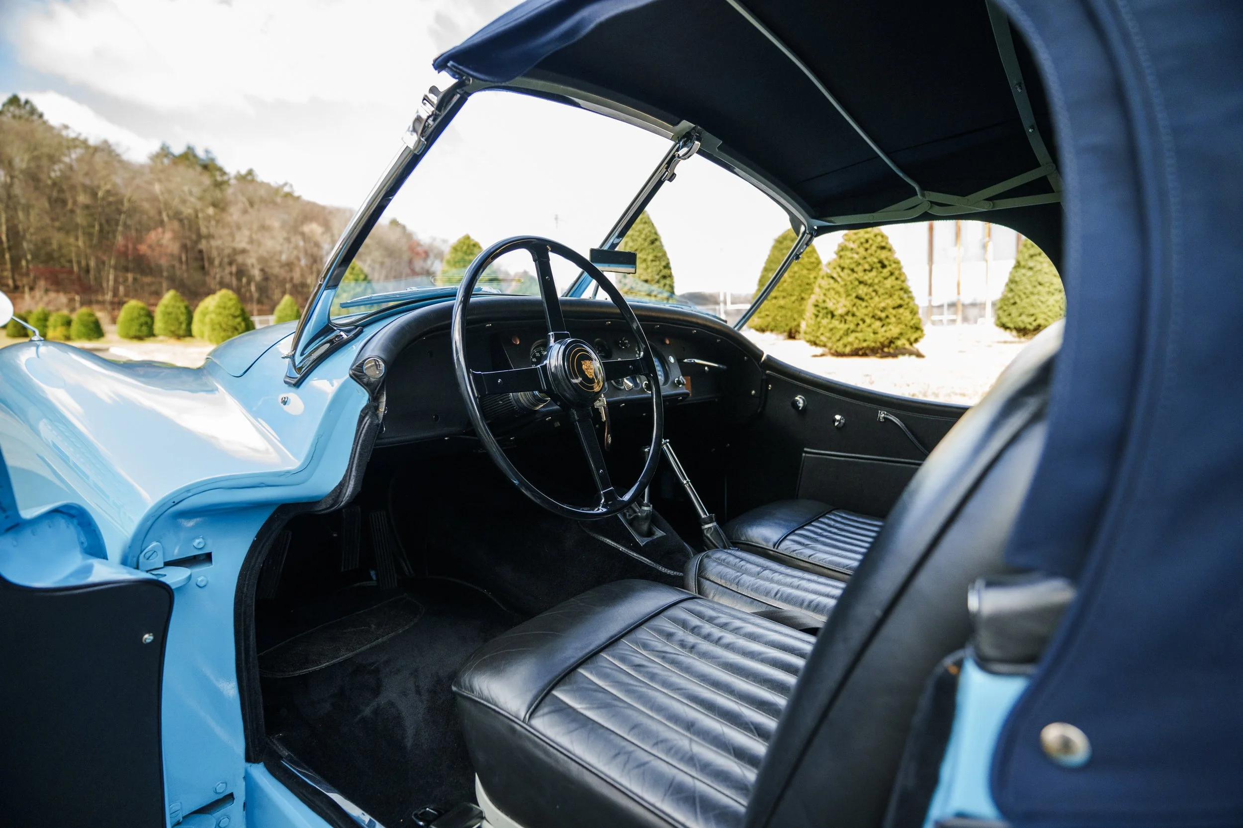 Interior of a vintage blue car showing the steering wheel, black leather seats, and dashboard details, with trees and blue sky visible outside