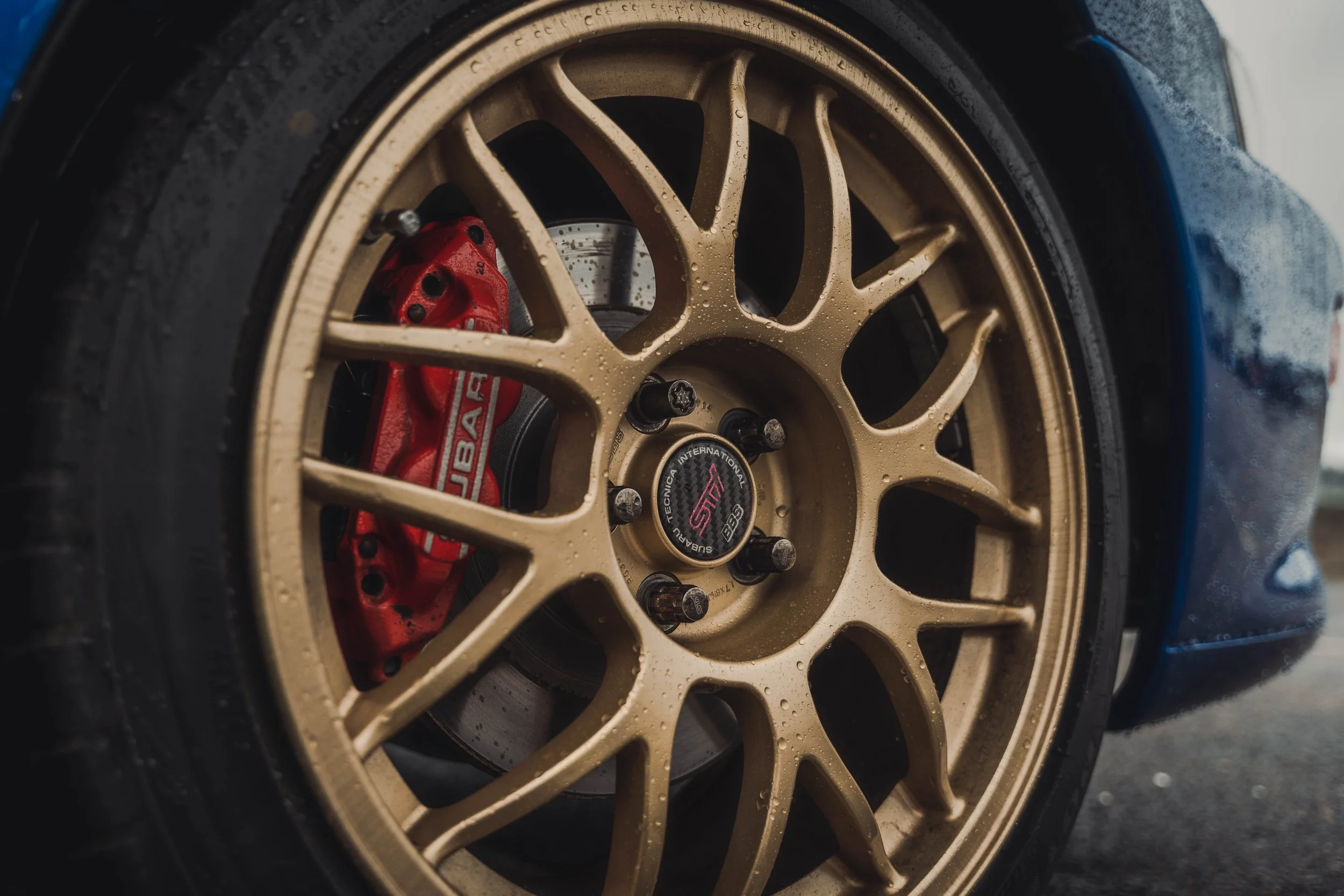 Close-up of a car wheel with a gold rim, red brake caliper, and a black tire, with water droplets on the rim.