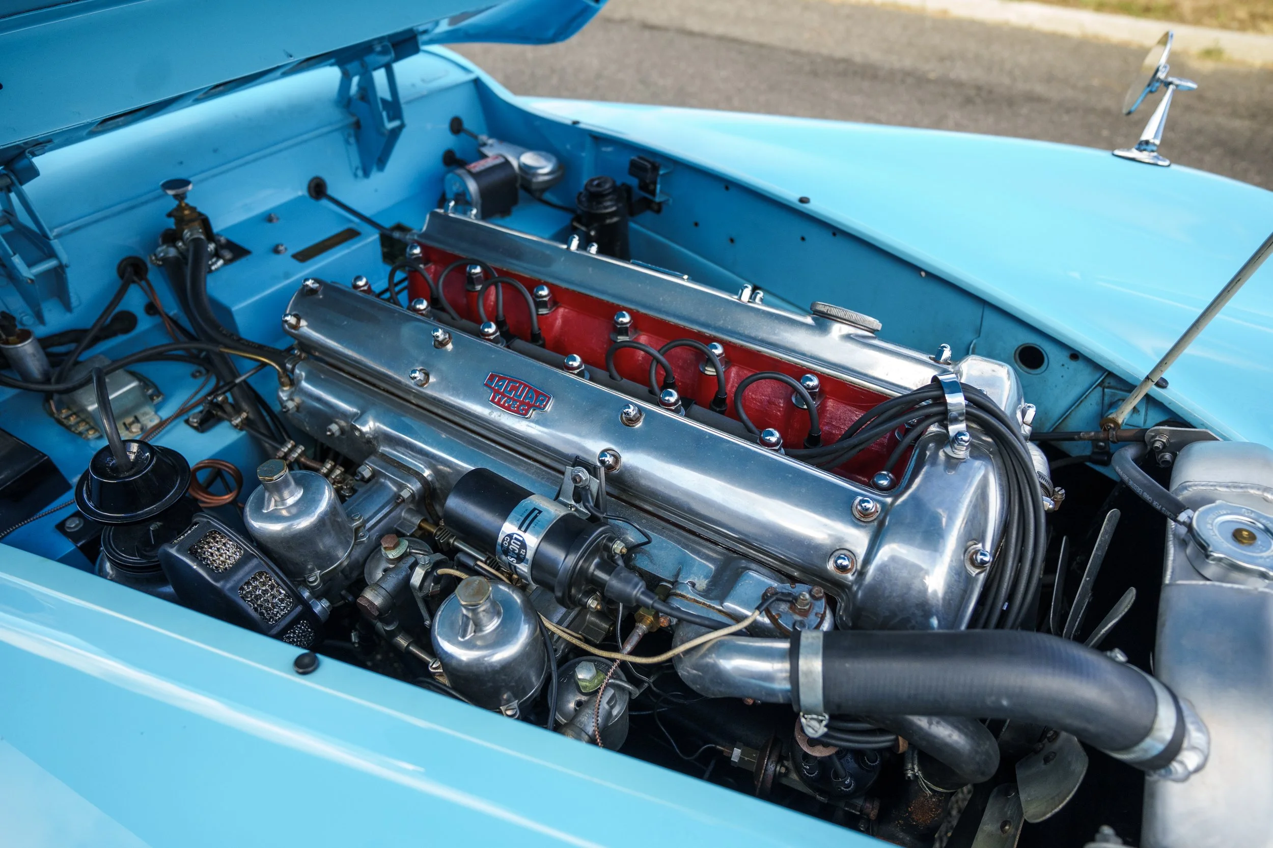 Close-up of a vintage car engine in a light blue sports car, with chrome components and a red label on the valve cover.