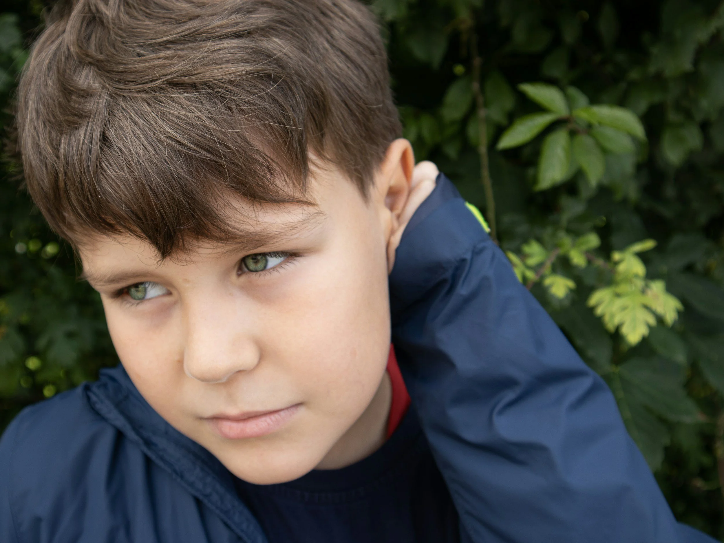 A young boy with brown hair and green eyes, wearing a navy blue jacket and red shirt, looking to the left, outdoors with green leaves in the background.
