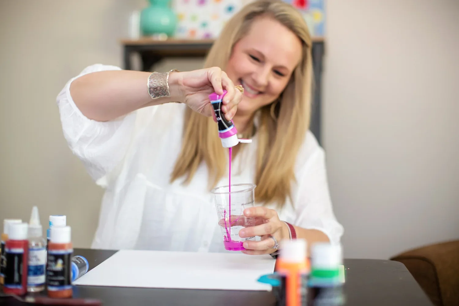 A woman is pouring pink paint into a plastic cup, surrounded by assorted bottles of paint on a table.