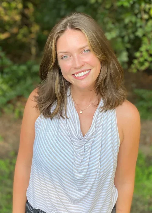 A smiling woman with shoulder-length light brown hair and blue eyes, wearing a sleeveless light blue and white striped top, standing outdoors with green foliage in the background.