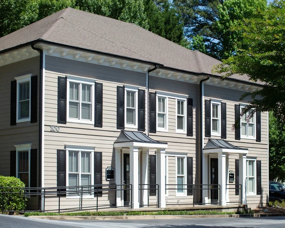 A two-story beige duplex with black shutters and white trim, featuring small covered entries supported by white columns, surrounded by greenery and trees.