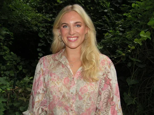 A smiling woman with long blonde hair wearing a pink and white floral shirt, standing outdoors in front of green foliage.