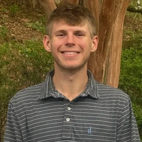 A young man smiling outdoors in front of a tree with green leaves, wearing a dark striped polo shirt.