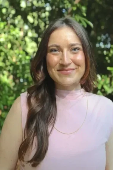 A young woman with long, wavy brown hair smiling outdoors with greenery in the background.