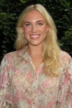 A woman with blonde hair wearing a floral blouse, smiling outdoors with a green leafy background.