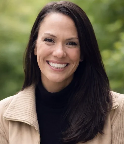 A woman with long dark hair smiling outdoors with a green blurred background.