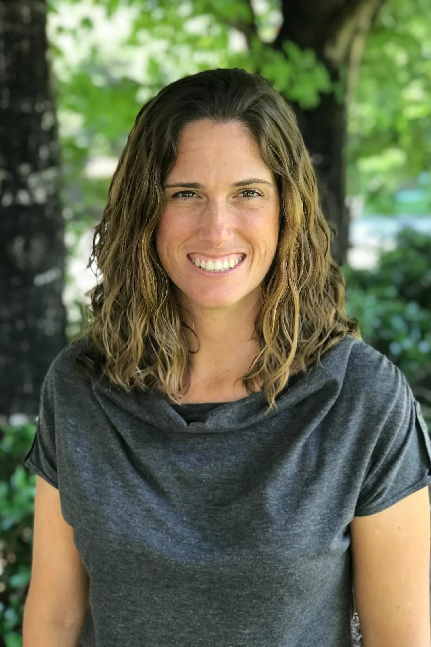 A woman with wavy brown hair and light skin, smiling, wearing a dark gray t-shirt, outdoors with green foliage in the background.