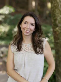 A woman standing outdoors in front of trees, smiling at the camera, wearing a sleeveless white top.