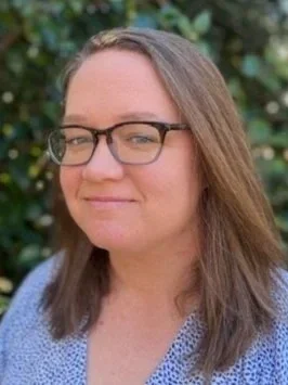 A woman with glasses and long brown hair smiling outdoors with green foliage in the background.