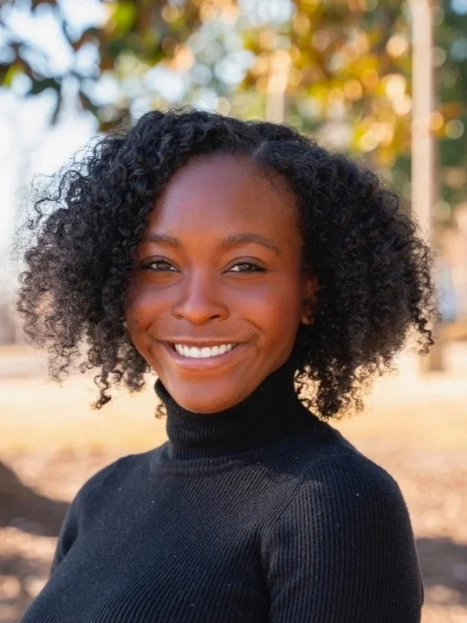 A smiling African American woman with curly black hair, wearing a black turtleneck sweater, outdoors with trees and sunlight in the background.