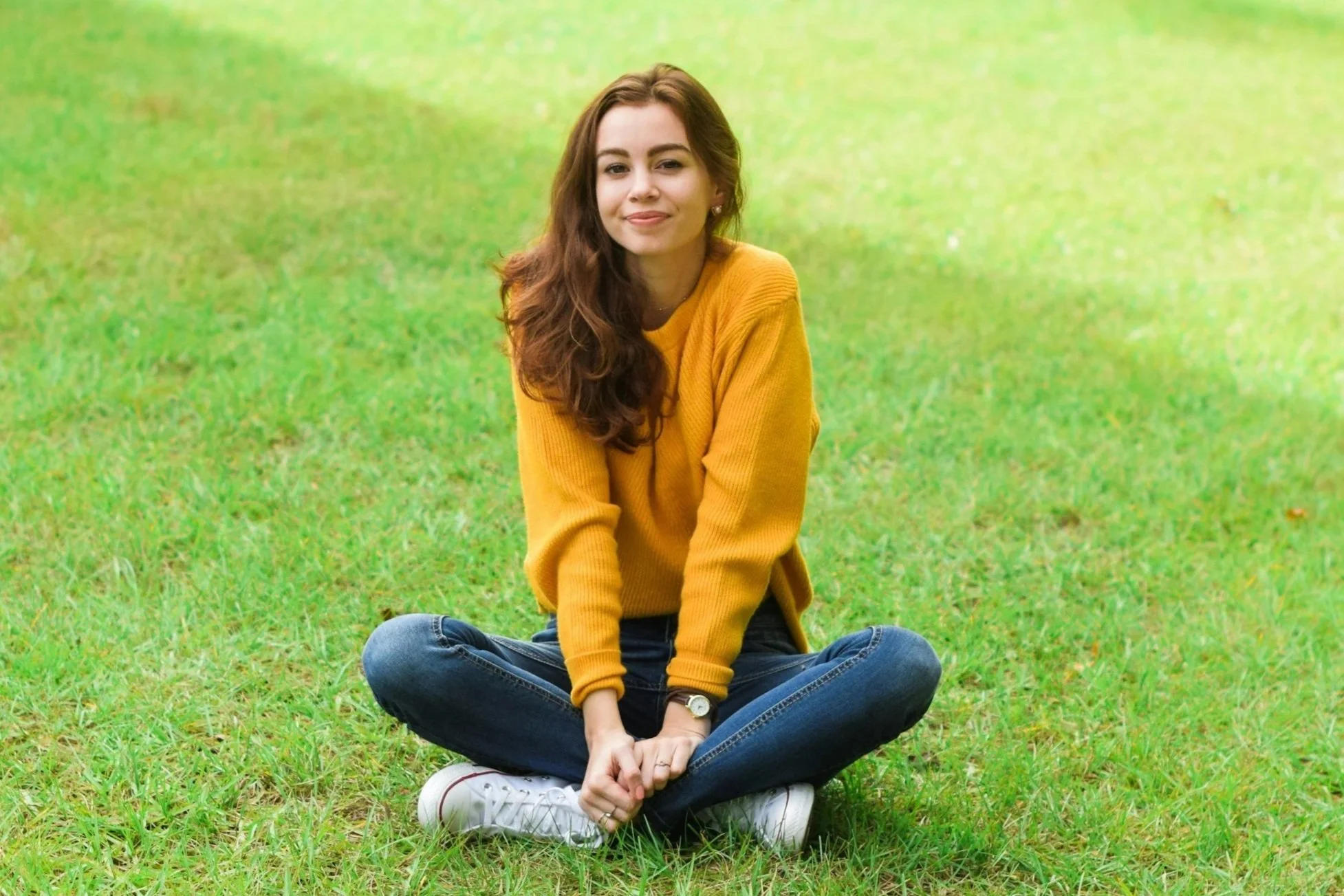 A young woman sitting cross-legged on green grass in a park, wearing a yellow sweater, blue jeans, and white sneakers, with long wavy brown hair, smiling slightly, looking at the camera.