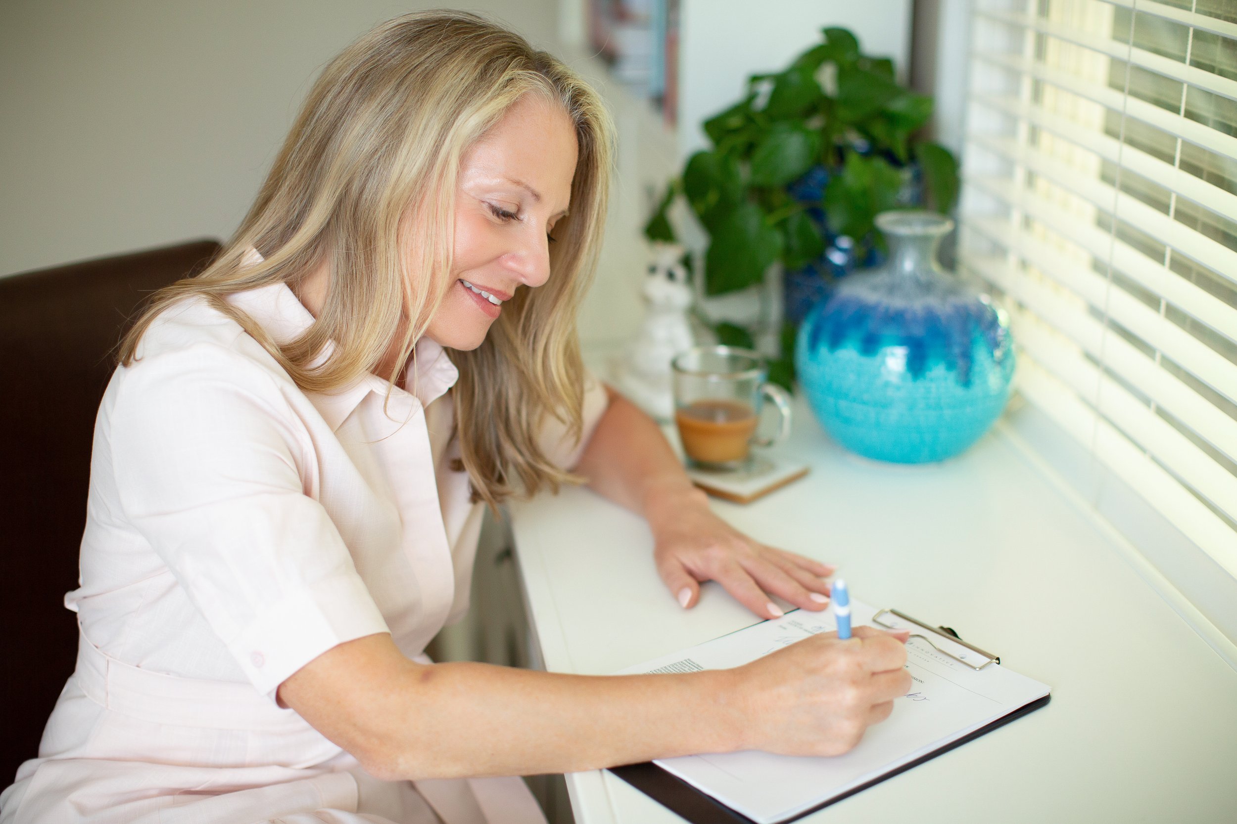 A woman with blonde hair smiling while writing in a notebook at a white table near a window with blinds. There are blue and white vases and a green plant in the background.